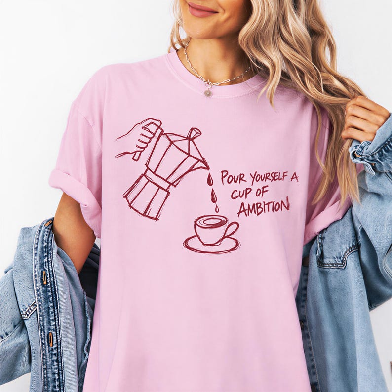 A woman wearing a pink t-shirt with a graphic design of a coffee pot pouring coffee into a cup, accompanied by the text "Pour yourself a cup of ambition".