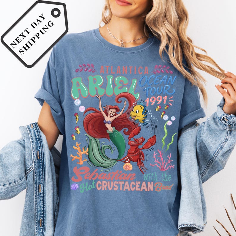 A woman wearing a blue t-shirt with a graphic design featuring the mermaid Ariel and the character Sebastian from the animated film "The Little Mermaid".
