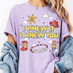 A person wearing a purple t-shirt with a cartoon-style design featuring a cowboy hat, star, clouds, and text.