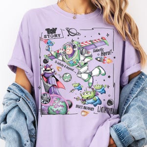 A person wearing a purple t-shirt with a graphic design featuring characters from the Toy Story franchise, including Buzz Lightyear and a group of smaller alien characters.