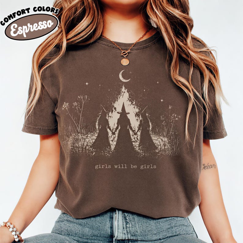 A woman wearing a brown t-shirt with a graphic design of three silhouetted figures and the text "girls will be girls" printed on it.