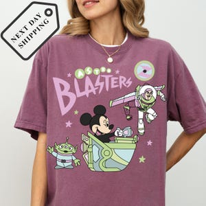 Pu&ograve; includere: T-shirt color malva con una grafica a cartoni animati di Topolino, Buzz Lightyear e un alieno. Il testo "Astro Blasters" &egrave; stampato sul davanti. La maglietta ha scollo tondo e maniche corte.