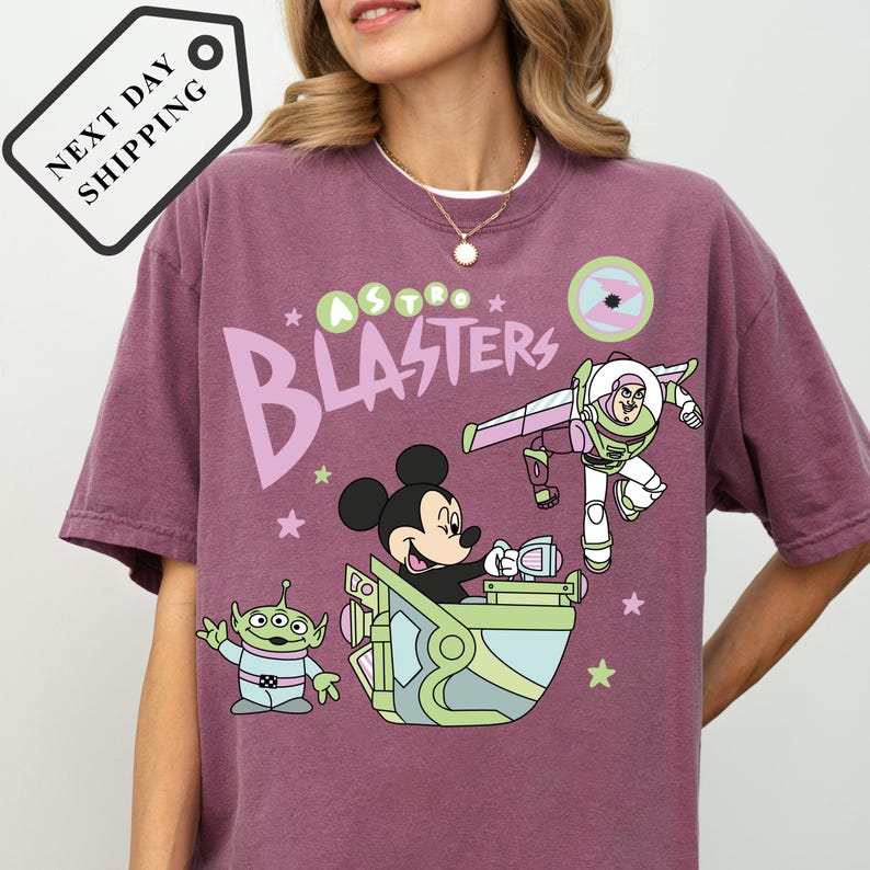 Pu&ograve; includere: T-shirt color malva con una grafica a cartoni animati di Topolino, Buzz Lightyear e un alieno. Il testo "Astro Blasters" &egrave; stampato sul davanti. La maglietta ha scollo tondo e maniche corte.