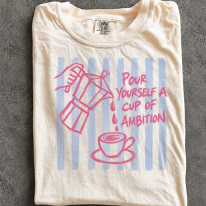 A white t-shirt with a graphic design depicting a coffee cup and a coffee pot, along with the text "Pour Yourself a Cup of Ambition".