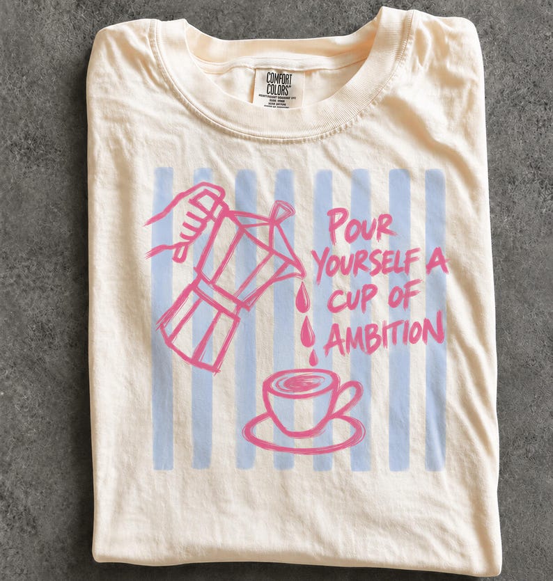 A white t-shirt with a graphic design depicting a coffee cup and a coffee pot, along with the text "Pour Yourself a Cup of Ambition".