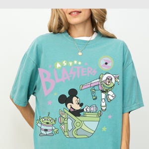 Pu&ograve; includere: T-shirt turchese con una grafica a cartoni animati di Topolino, Buzz Lightyear e un alieno. Le parole "Astro Blasters" sono visualizzate in un carattere grande e stilizzato. Il design include stelle e una grafica circolare.