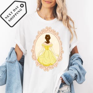 A woman wearing a white t-shirt with a graphic of a woman in a yellow dress, standing in a decorative frame.