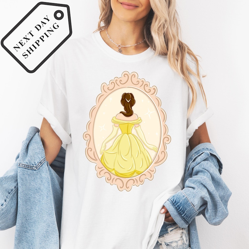 A woman wearing a white t-shirt with a graphic of a woman in a yellow dress, standing in a decorative frame.