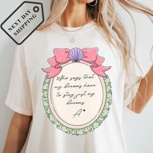 A white t-shirt with a graphic design featuring a pink bow and a text quote about pursuing one's dreams.