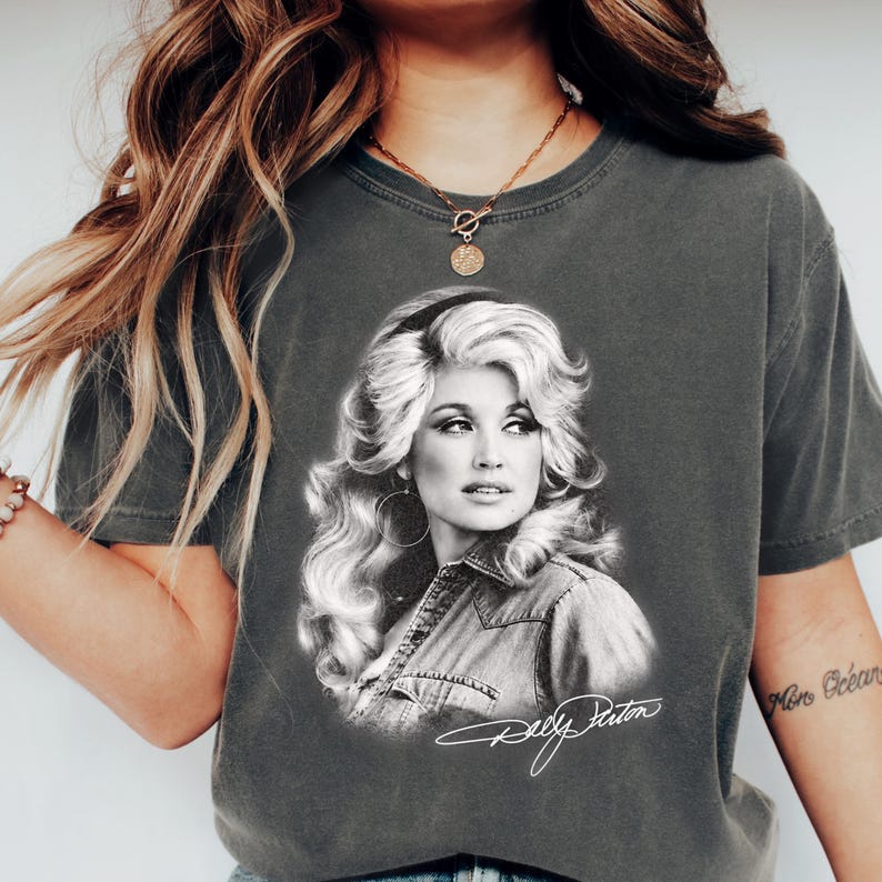 May include: A charcoal gray short-sleeve t-shirt featuring a black and white photographic print of Dolly Parton with her signature voluminous blonde hair. The graphic includes a white cursive signature of Dolly Parton printed below the portrait.
