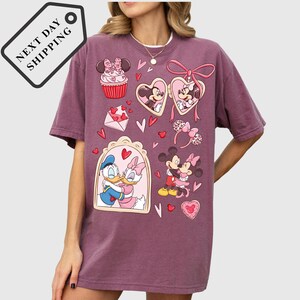 A woman wearing a purple t-shirt with a graphic design featuring Disney characters such as Minnie Mouse, Donald Duck, and Mickey Mouse, along with various heart-shaped and heart-studded elements.