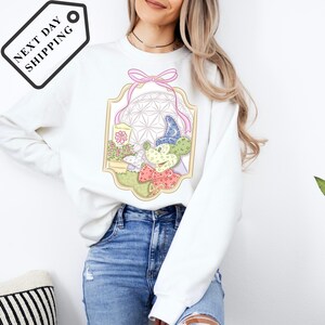 A woman wearing a white sweatshirt with a colorful graphic design on it, standing against a white background.