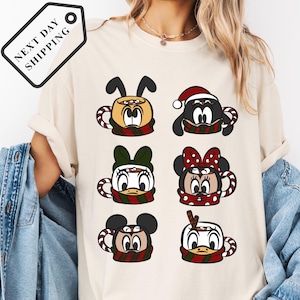 May include: Cream-colored t-shirt featuring six cartoon character mugs with festive scarves and hats. The mugs depict various characters, including Mickey and Minnie Mouse, and Donald Duck. The tag on the top left says "NEXT DAY SHIPPING".