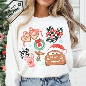 A woman wearing a white long-sleeved shirt with a Christmas-themed design featuring various holiday-related icons and characters, such as a Santa hat, a gingerbread man, and a car.