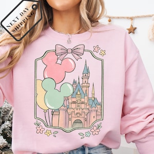 May include: A light pink sweatshirt featuring a whimsical castle design with Mickey Mouse balloons in pastel colors. The design is framed by a decorative border with a bow and floral accents. The tag reads "NEXT DAY SHIPPING".