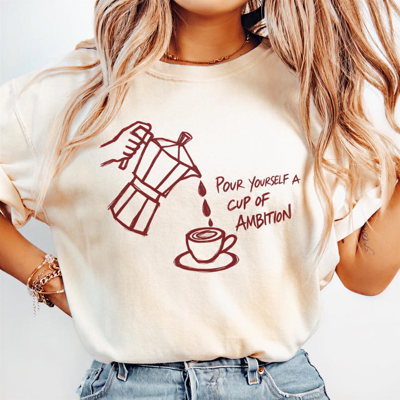 A woman wearing a white t-shirt with a graphic design of a coffee pot pouring coffee into a cup, accompanied by the text "Pour Yourself a cup of Ambition".