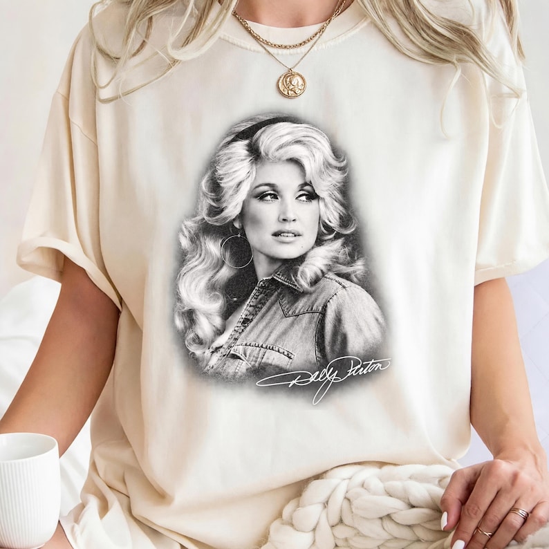 May include: A cream-colored t-shirt featuring a black and white graphic print of Dolly Parton with her signature voluminous hair and a denim shirt. The design includes a stylized signature of Dolly Parton in white script at the bottom of the image.