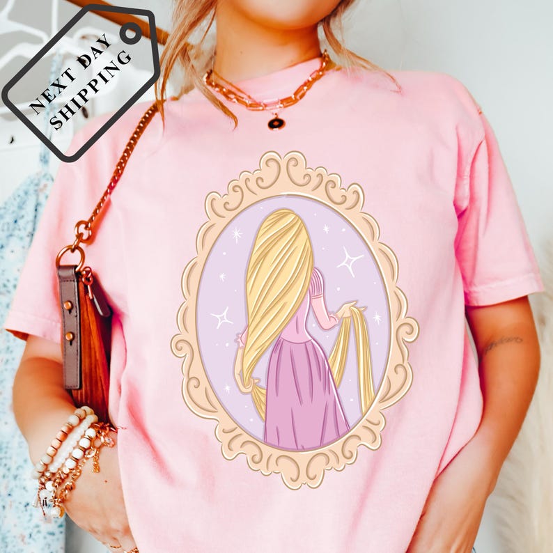 A woman wearing a pink t-shirt with a design of a blonde woman in a pink dress, standing in a frame with stars.