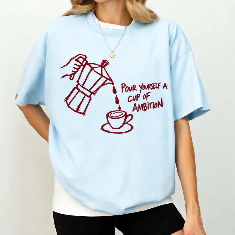 A woman wearing a light blue t-shirt with a graphic design depicting a coffee pot pouring coffee into a cup, accompanied by the text "Pour yourself a cup of ambition".