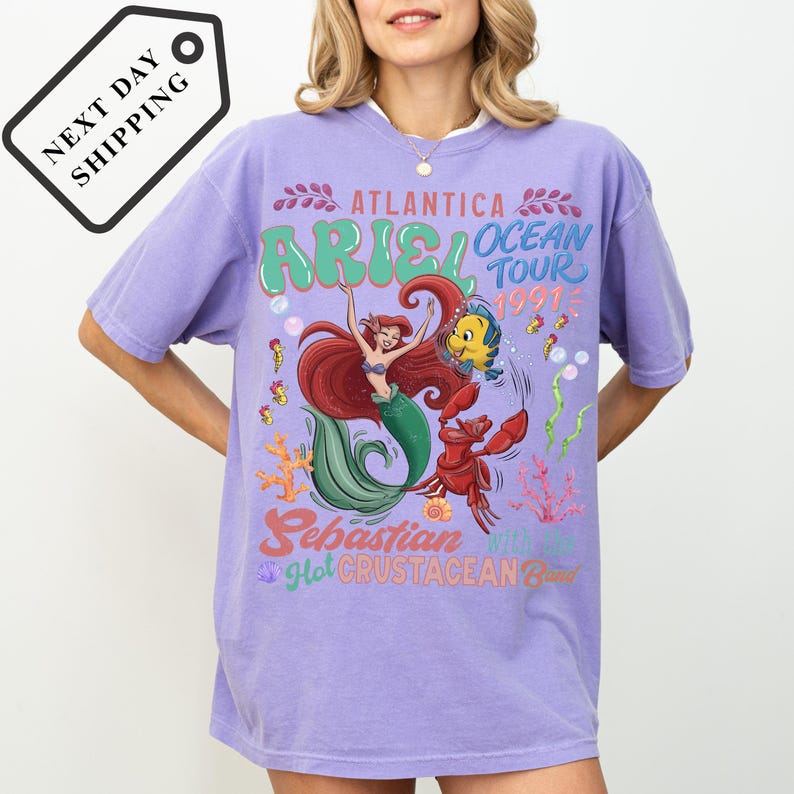 A woman wearing a purple t-shirt with a colorful graphic design featuring the mermaid Ariel and the crab Sebastian from the Disney film "The Little Mermaid".