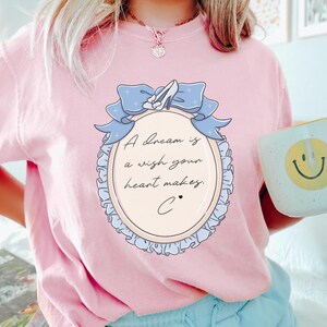 A woman wearing a pink t-shirt with a graphic design and text, holding a coffee mug with a smiley face.