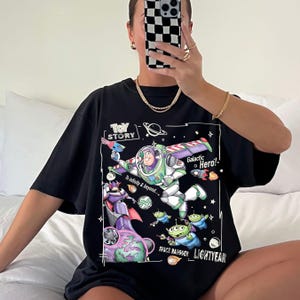 A person is sitting on a bed and taking a selfie with a black t-shirt featuring a graphic of a character from the Toy Story movie.