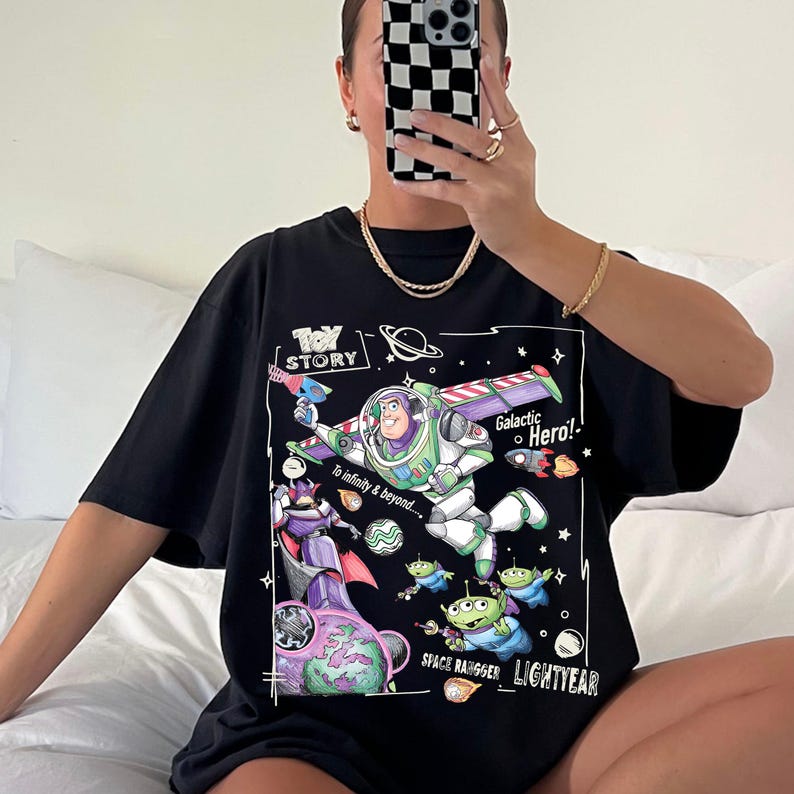 A person is sitting on a bed and taking a selfie with a black t-shirt featuring a graphic of a character from the Toy Story movie.
