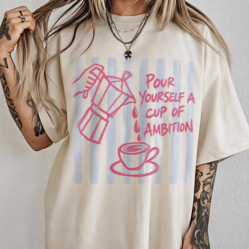 A person wearing a white t-shirt with a graphic design of a coffee cup pouring a cup of coffee, accompanied by the text "Pour Yourself a Cup of Ambition".