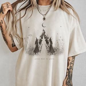 A person wearing a white t-shirt with a graphic design featuring three women with long hair and a crescent moon, along with the text "girls will be girls".