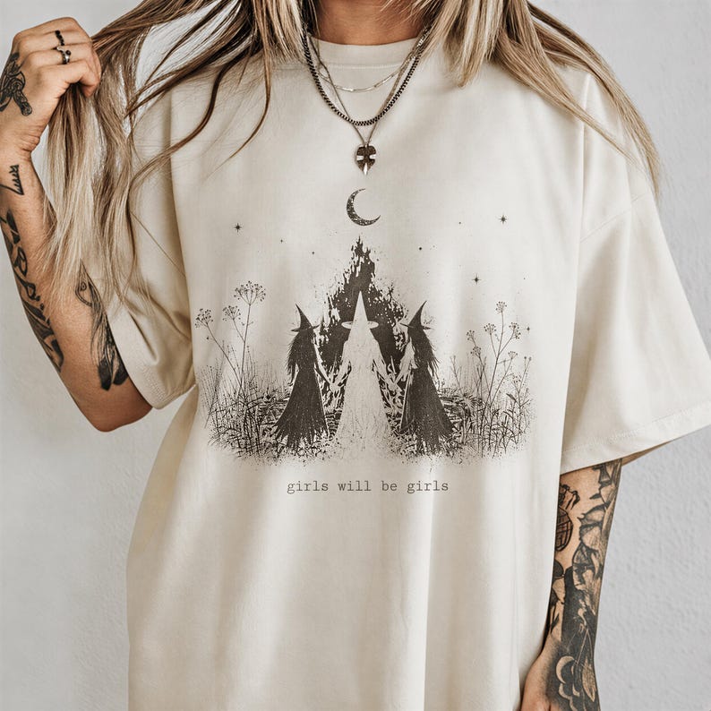 A person wearing a white t-shirt with a graphic design featuring three women with long hair and a crescent moon, along with the text "girls will be girls".