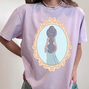 A woman wearing a purple t-shirt with a graphic of a woman in a mirror frame.