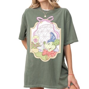 A woman wearing a green t-shirt with a colorful graphic design featuring a cartoon character and various decorative elements.