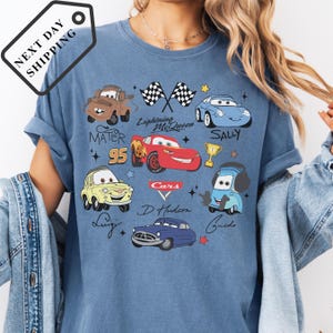 A person wearing a blue t-shirt with various cartoon car characters and designs, including Lightning McQueen, Sally, Mater, and other popular Disney characters.