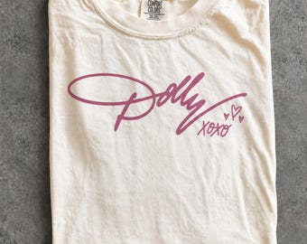 May include: A cream-colored short-sleeve t-shirt featuring the word Dolly in a dark pink cursive script across the chest. Below the name, the text XOXO is written in a matching pink script, accompanied by three small hand-drawn heart outlines.