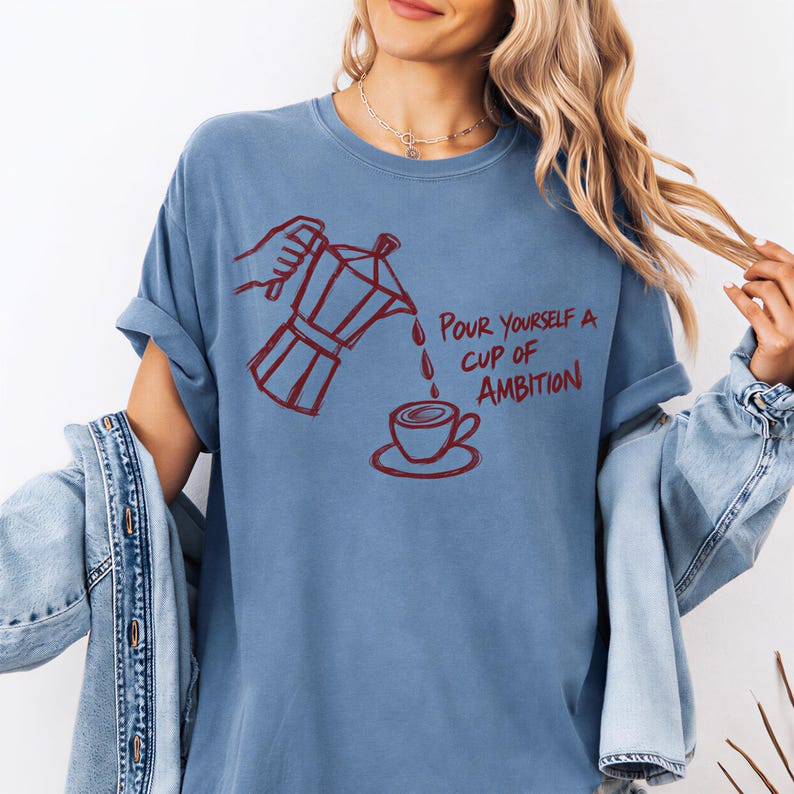 A woman wearing a blue t-shirt with a graphic design of a coffee pot pouring coffee into a cup, accompanied by the text "Pour yourself a cup of ambition".