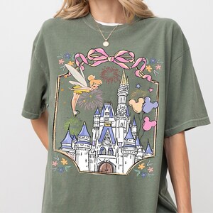 A green t-shirt with a graphic design depicting a castle surrounded by colorful flowers and a fairy.