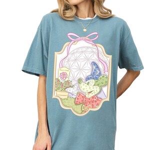 A woman wearing a blue t-shirt with a colorful graphic design featuring a flower, a bow, and various other decorative elements.