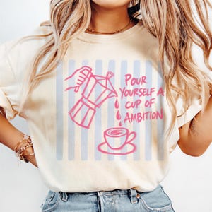 A woman wearing a white t-shirt with a graphic design of a coffee cup and the text "Pour yourself a cup of ambition" printed on it.