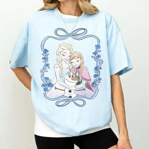 May include: A light blue t-shirt featuring a graphic print of two blonde-haired characters and a snowman inside a decorative blue floral frame with bows at the top and bottom. The characters are embracing the snowman in a central illustration.