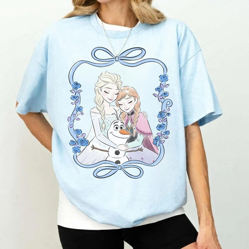 May include: A light blue t-shirt featuring a graphic print of two blonde-haired characters and a snowman inside a decorative blue floral frame with bows at the top and bottom. The characters are embracing the snowman in a central illustration.