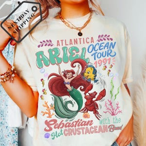 A person wearing a white t-shirt with a colorful graphic design featuring the character Ariel from The Little Mermaid and the band Sebastian from The Hot Crustacean Band.