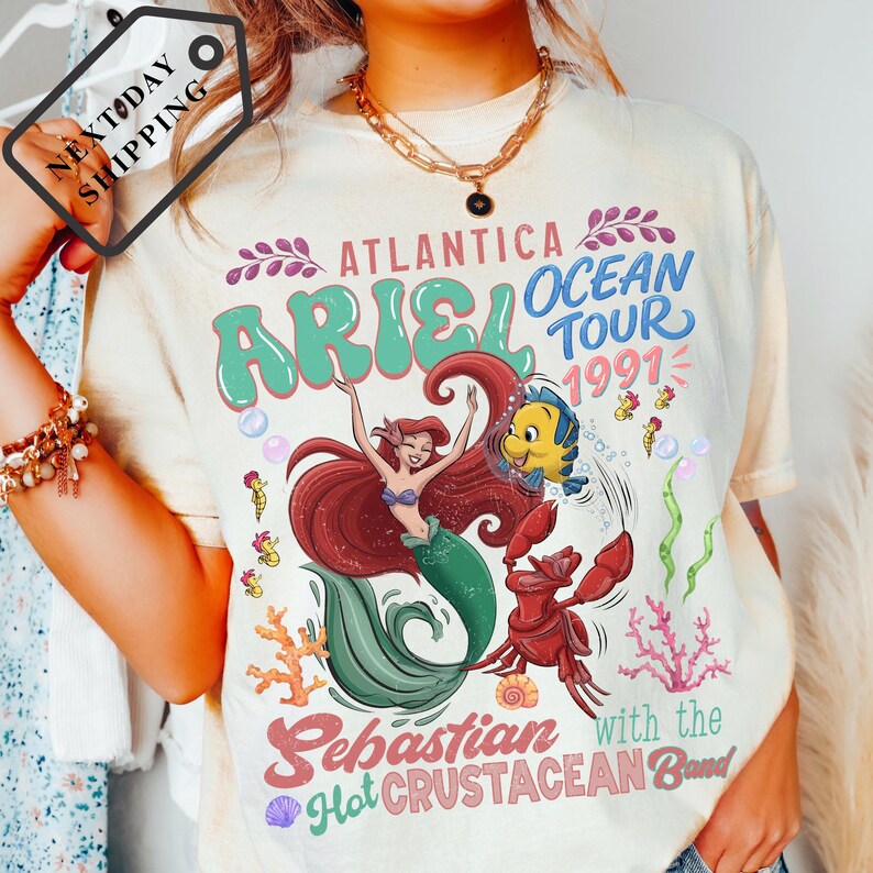 A person wearing a white t-shirt with a colorful graphic design featuring the character Ariel from The Little Mermaid and the band Sebastian from The Hot Crustacean Band.