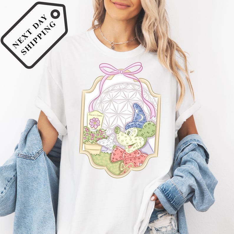 A woman wearing a white t-shirt with a colorful graphic design of a space station and various objects, including a flower and a cactus, is standing against a white background.