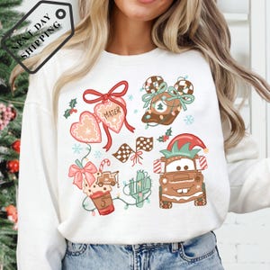 A woman wearing a white long-sleeved shirt with a Christmas-themed graphic design featuring various holiday-related elements such as a Santa hat, a gingerbread man, a reindeer, and a Christmas tree.