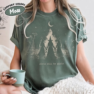 A woman wearing a green t-shirt with a graphic design of a forest and a crescent moon, holding a teacup.