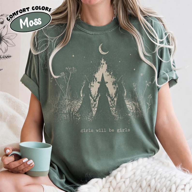 A woman wearing a green t-shirt with a graphic design of a forest and a crescent moon, holding a teacup.