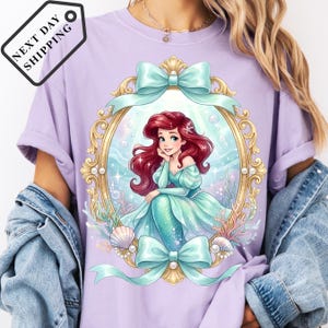 A person wearing a purple t-shirt with a graphic of a mermaid character, surrounded by a decorative frame and ribbons.