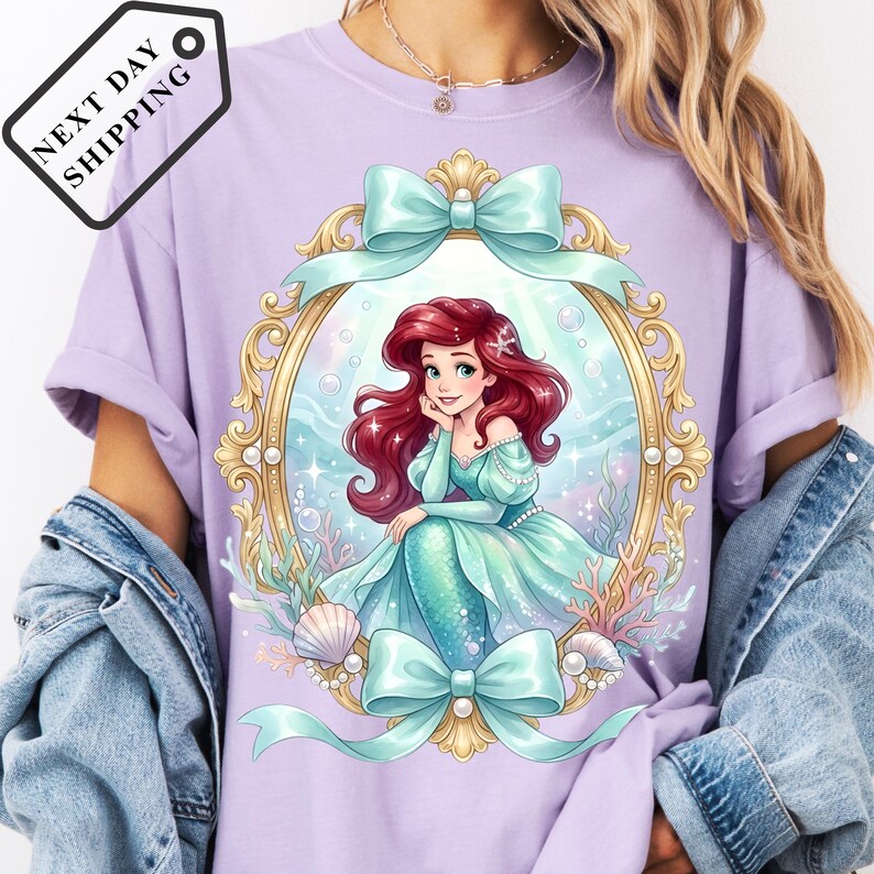 A person wearing a purple t-shirt with a graphic of a mermaid character, surrounded by a decorative frame and ribbons.