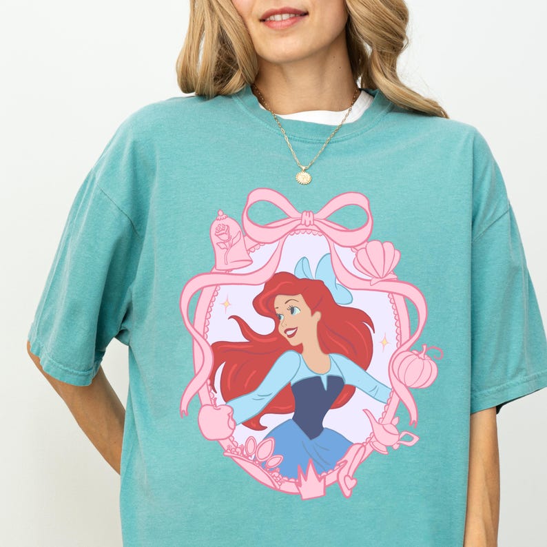A woman wearing a turquoise t-shirt with a graphic of a young girl with red hair and a blue dress, surrounded by pink ribbons and other decorative elements.