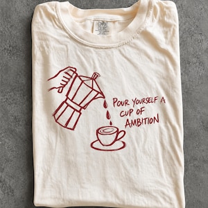 A white t-shirt with a red graphic design depicting a coffee pot pouring coffee into a cup, accompanied by the text "Pour yourself a cup of ambition".